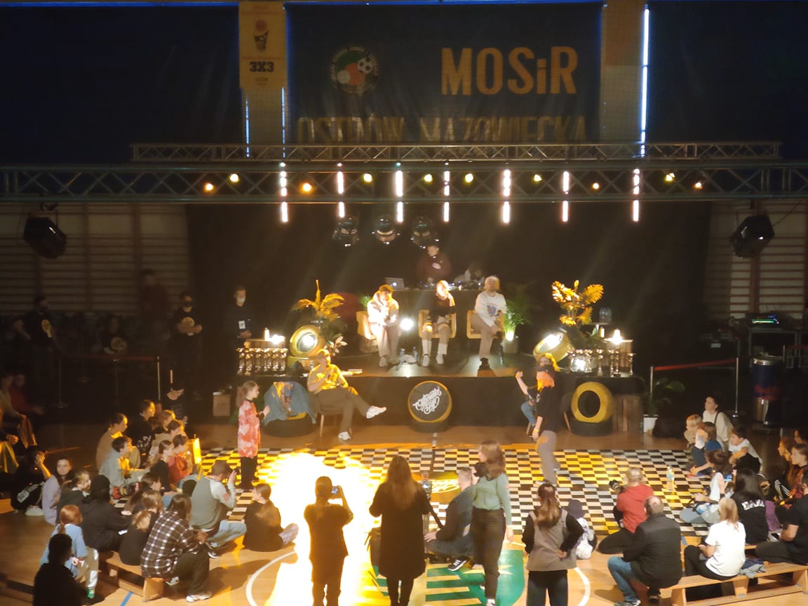 Występ artystyczny na scenie w hali MOSiR Ostrów Mazowiecka, z muzykami i dziecięcą publicznością siedzącą na podłodze. Widoczny baner MOSiR.