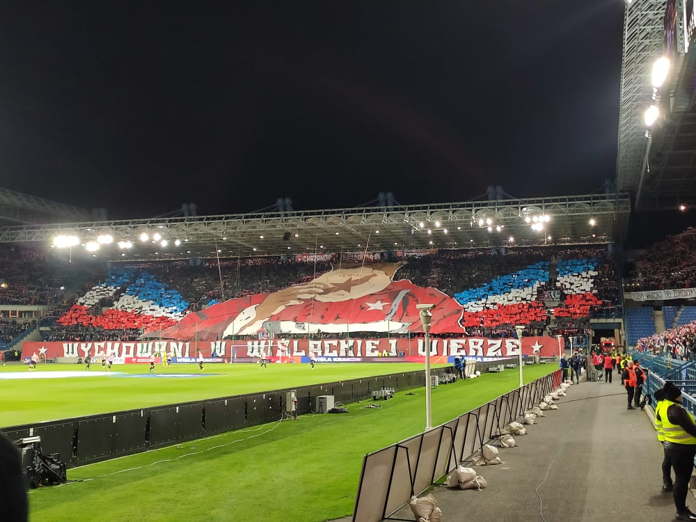 Trybuny stadionu piłkarskiego wypełnione kibicami z efektowną sektorówką w barwach czerwono-niebiesko-białych oraz napisem 'WYCHOWANI W WIŚLACKIEJ WIERZE'.